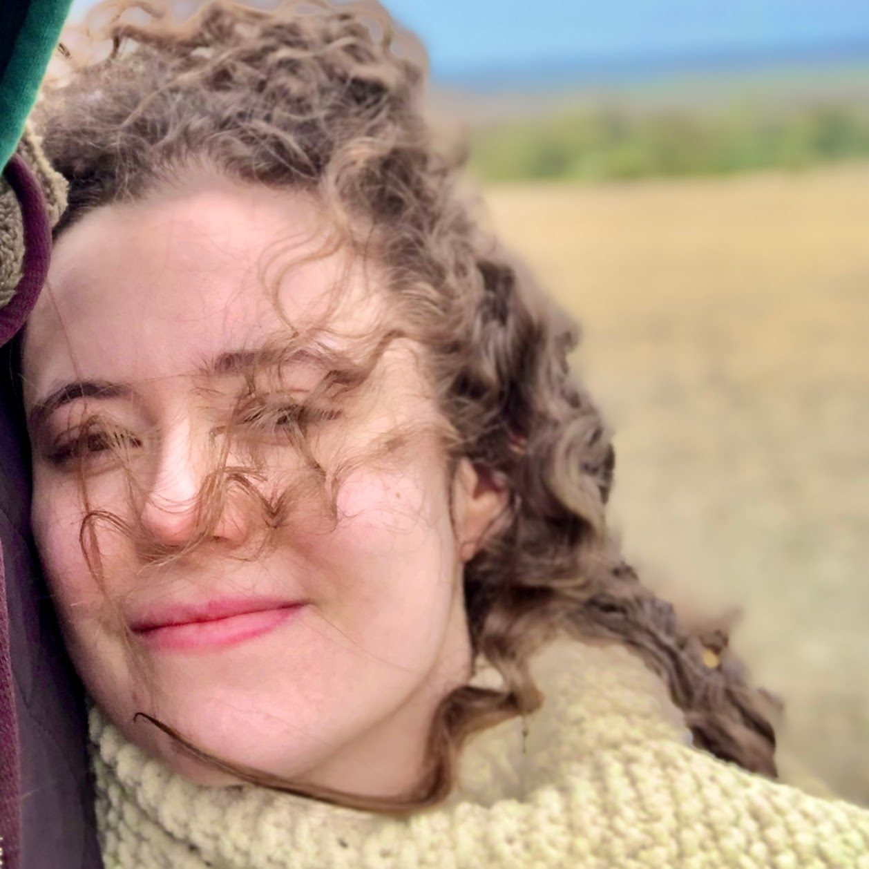Woman with curly brown hair and yellow scarf smiling with countryside in the background